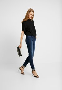 Černý top s krátkým rukávem v kombinaci s tmavě modrými skinny džíny. Modelka má na sobě černé sandály na vysokém podpatku a drží malou černou kabelku se řetězovými detaily.