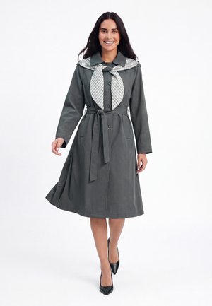 Donna sorridente mentre cammina, indossa un trench coat scuro con cintura e una sciarpa a quadri legata intorno al collo con tacchi neri.