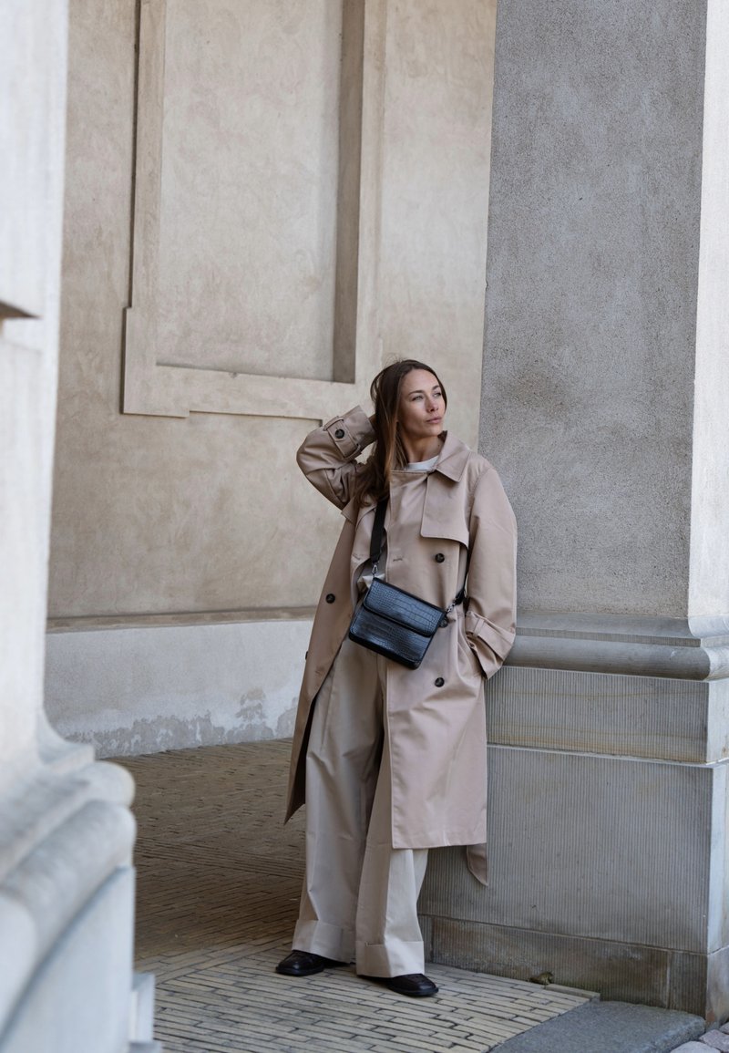 Femme appuyée contre une colonne, portant un trench-coat beige, un pantalon large, des chaussures noires et un sac bandoulière noir, regardant sur le côté.