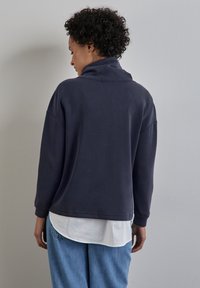 Felpa blu scuro con colletto alto, spalle scese e maniche lunghe. Indossata sopra una camicia bianca, con una vestibilità comoda e una texture morbida.