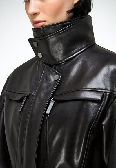 Záber zblízka na osobu nosiacu čiernu koženú bundu s vysokými golierom, patentkami a na zips uzatvárateľnými náprsné vrecká.