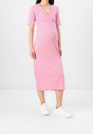 Kobieta o ciemnych włosach, ubrana w różową prążkowaną dzianinową sukienkę midi i białe trampki, stojąca na jednolitym jasnym tle.