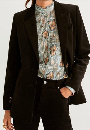 Femme portant un blazer et un pantalon en velours côtelé brun foncé avec un chemisier plissé à col montant à motifs floraux bleu, orange et beige.