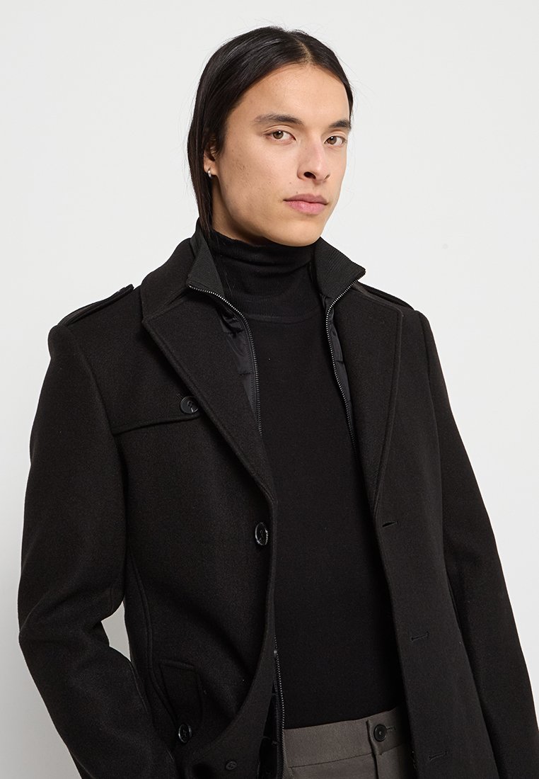 Schwarzer Wollmischmantel mit Kragen und doppelter Knopfleiste. Hat eine glatte Textur und eine strukturierte Silhouette, die über einem schwarzen Rollkragenpullover getragen wird.