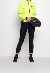 Klar gul cykeljacka med reflekterande detaljer, matchad med svarta leggings. Modellen håller en svart hjälm och visar upp sportutrustning.