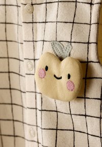 Fruit beige en peluche avec joues roses et sourire cousu sur tissu blanc à motif quadrillé noir avec boutons visibles.