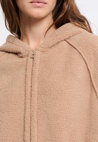 Felpa in pile beige con chiusura a zip e superficie testurizzata. Presenta un cappuccio e maniche raglan, evidenziando un design accogliente.