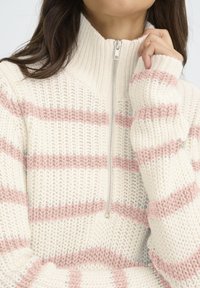 Jersey de punto con un alto cuello, cierre de cremallera y rayas horizontales rosas sobre un fondo crema. Textura suave y corte holgado.