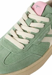 Baskets en daim vert avec des lacets crème, des accents texturés et une languette rose arborant un logo métallique. Semelle en caoutchouc, design athlétique classique.