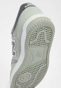 Baskets grises avec une semelle en caoutchouc, motifs texturés sur la semelle extérieure, et un accent noir au talon avec le logo New Balance.