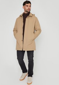 Beige waterdichte jas met capuchon, drukknoopsluiting en zijzakken, gedragen over een donkerbruine top en gecombineerd met donkerdenim en witte sneakers.