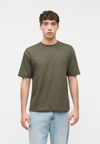 Νέος άνδρας με κοντά καστανά, σγουρά μαλλιά, ντυμένος με κοντομάνικο ελιάς πράσινο μπλουζάκι και ανοιχτό μπλε τζιν, στέκεται μπροστά σε ένα απλό λευκό φόντο.