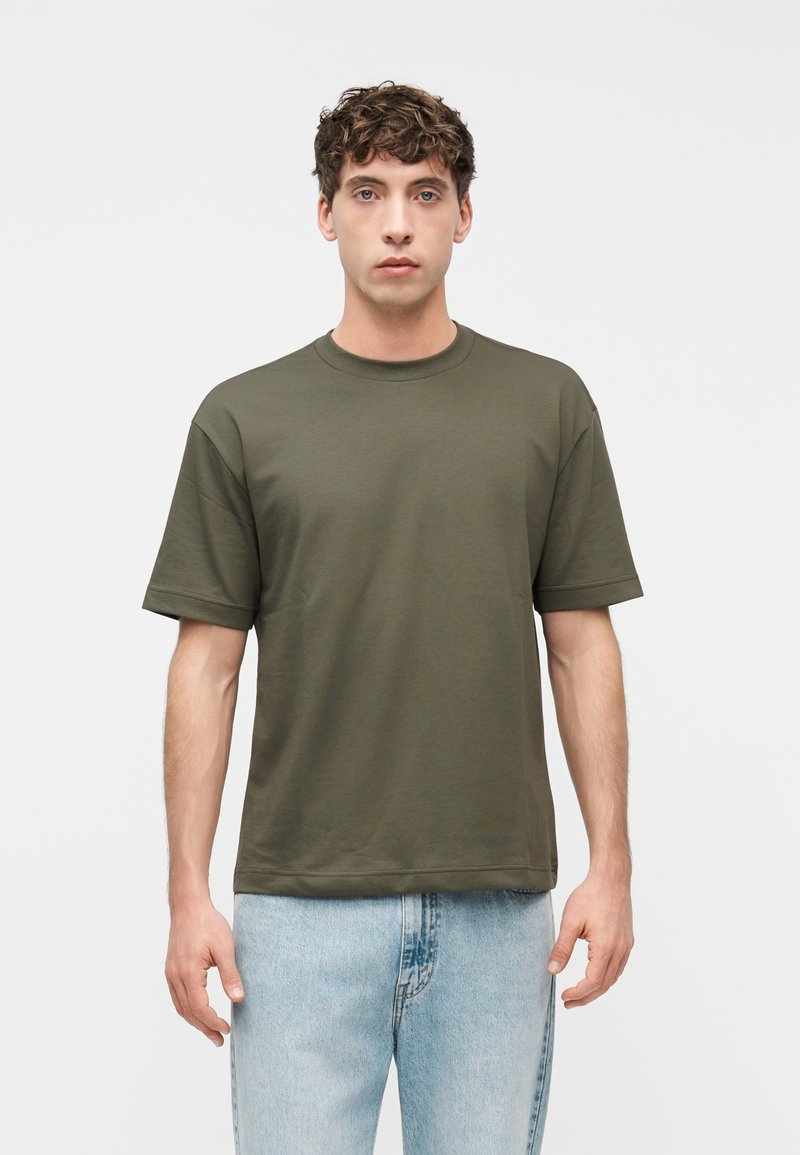 Νέος άνδρας με κοντά καστανά, σγουρά μαλλιά, ντυμένος με κοντομάνικο ελιάς πράσινο μπλουζάκι και ανοιχτό μπλε τζιν, στέκεται μπροστά σε ένα απλό λευκό φόντο.