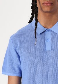 Lyseblå strikket polo skjorte med korte ermer, klassisk krage og to frontknapper. Teksturen er luftig og pustende.