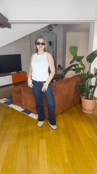 Hvit ermeløs topp, mørkeblå jeans og beige fottøy. Rom med tregulv, brun sofa, grønn plante og TV-enhet.