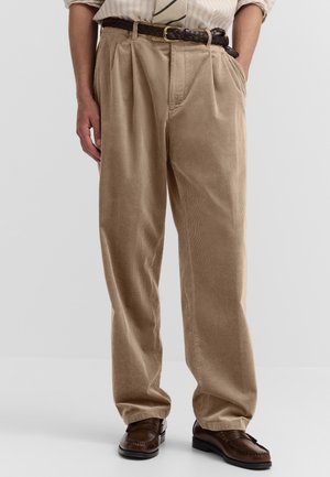 Beige Cordhose, getragen mit einem braunen geflochtenen Gürtel, kombiniert mit braunen Lederloafers, gezeigt von der Taille bis zu den Füßen.
