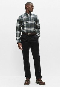 Koszula w kratę zapinana na guziki w ciemnozielono-białym kolorze, zestawiona z ciemnymi sztruksowymi spodniami i brązowymi skórzanymi butami. Model stoi z rękami w kieszeniach.