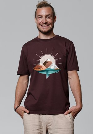 T-shirt en coton bordeaux avec un graphique de montagnes et d'eau en orange et en teal, design rond avec des rayons de soleil. Coupe décontractée, manches courtes.