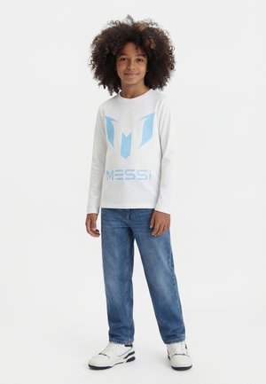 Dieťa s kučeravými vlasmi, oblečené v bielej dlhorukávovej košeli s nápisom Messi, modrých džínsoch a bielych teniskách, stojace pred bielym pozadím.