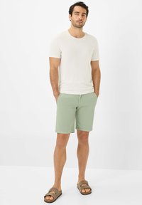 T-shirt color beige chiaro, pantaloni corti verde menta con una texture liscia e sandali marroni con design a doppia cinghia, in piedi davanti a uno sfondo semplice.