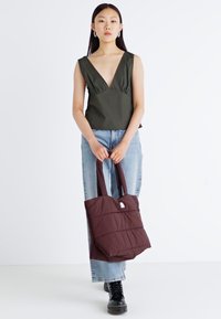 Braune, gesteppte Tragetasche mit zwei stabilen Griffen, präsentiert von einem Modell, das ein dunkelgrünes Oberteil und hellblaue Jeans trägt. Schlichter weißer Hintergrund.