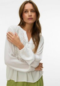 Femme aux longs cheveux bruns portant un chemisier blanc avec des détails en dentelle et un pantalon vert, posant les bras croisés devant un fond uni.