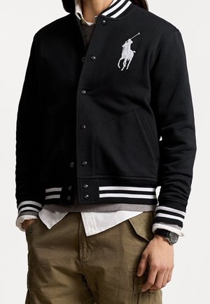Personne portant une veste universitaire noire avec des rayures blanches aux poignets et à l'ourlet, une chemise blanche, un pantalon cargo marron et une montre-bracelet.