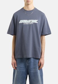 Szary bawełniany t-shirt o luźnym kroju, z białym logo AVIREX DEPT. na piersi i krótkimi rękawami. W połączeniu z jasnoniebieskimi dżinsami.