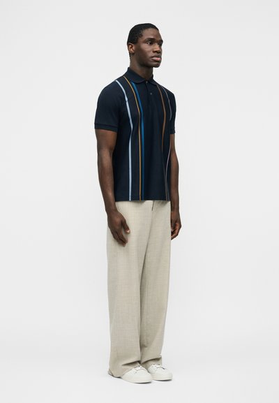 Homme debout, faisant face à droite, portant un polo noir à rayures verticales multicolores, un pantalon large beige et des baskets blanches sur un fond blanc.