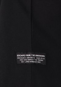 Tela negra con textura de canalé, que presenta una etiqueta texturizada que dice "ESCAPE DE / LO DESCONOCIDO" en texto blanco.