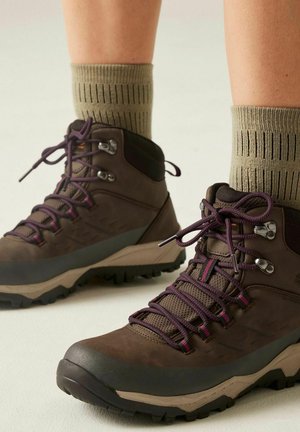 Brown hiking boots with purple laces worn with beige socks featuring vertical black stripes on a light background.