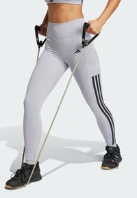 Leggings atléticos de color gris claro con acentos de tres rayas negras, combinados con zapatillas deportivas negras. El usuario sostiene bandas de resistencia con empuñaduras.