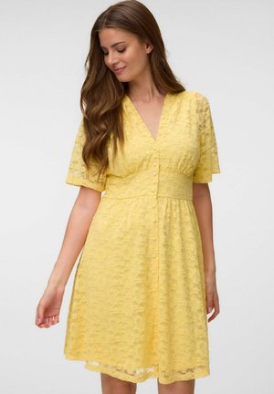 Donna con lunghi capelli castani che indossa un vestito giallo in pizzo fino al ginocchio con maniche corte e bottoni sul davanti, guarda verso il basso e sorride dolcemente.