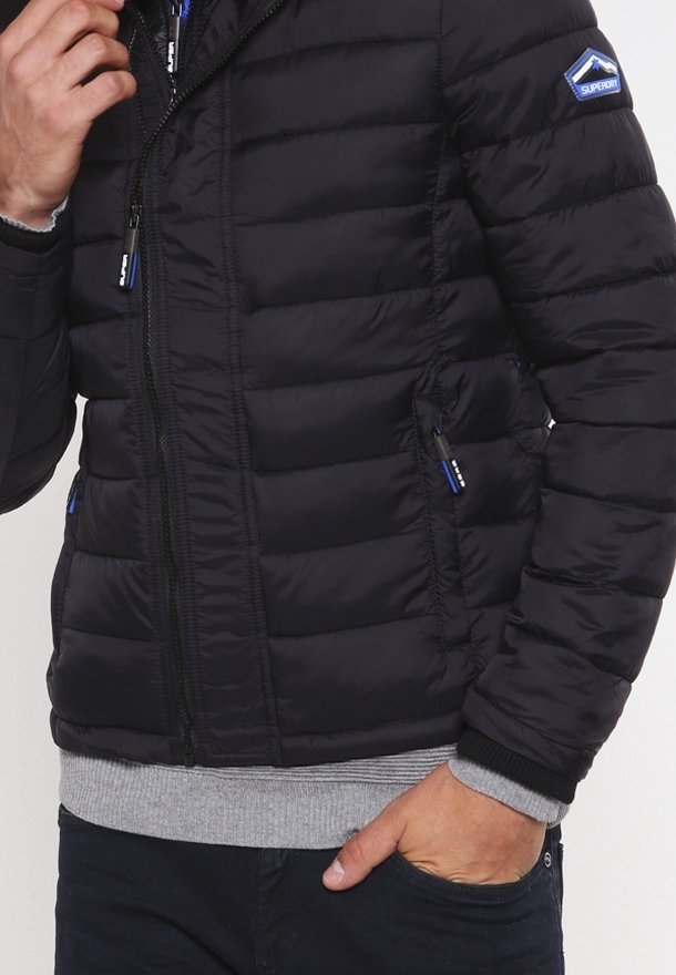 Sort puffer-jacket med quiltet tekstur, lynlåslukning foran, to sidelommer og grå strikkede manchetter. Har et blåt logo på ærmet.