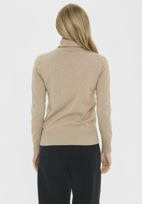 Beżowy sweter z golfem, z długimi rękawami, ściągaczami przy mankietach i u dołu, dopasowany krój. Noszony z ciemnymi, wysokimi spodniami, widok z tyłu.