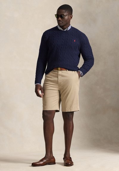 Άνδρας που φοράει μπλε πουλόβερ με πλεκτό σχέδιο, ριγέ πουκάμισο με γιακά, μπεζ σορτς, καφέ ζώνη και μοκασίνια, σκουρόχρωμα γυαλιά ηλίου, στέκεται σε ουδέτερο φόντο.