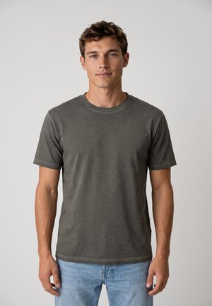 Joven con el pelo corto y castaño, vistiendo una camiseta lisa de color gris oscuro y vaqueros azul claro, de pie frente a un fondo claro y liso.