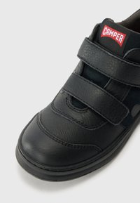 Čierne kožené topánky s okrúhlym prstom, dvoma suchými zipsami a červeno-bielym logom "Camper". Viditeľné sú textúrované a hladké povrchy.