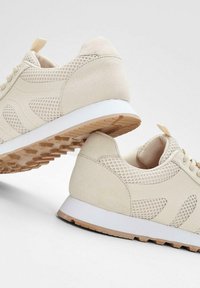 Scarpe atletiche beige con pannelli in rete e parte superiore in tessuto liscio. Suola in gomma con un motivo di battistrada testurizzato; accenti di design minimi.