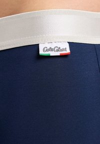 Marineblauw kledingstuk met een brede, gestructureerde witte elastische tailleband met een label waarop "Carlo Colucci" staat en een klein Italiaans vlagontwerp.