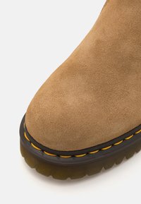 Tan suède enkellaars met ronde neus, zwarte rubberen zool en gele stiksels langs de rand. Het oppervlak lijkt zacht met een matte afwerking.