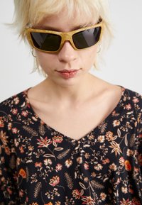 Lunettes de soleil avec une monture en écaille de tortue jaune et des verres foncés. Haut noir à motifs floraux avec décolleté en V et manches courtes.