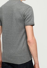 Mand iført en ensfarvet grå t-shirt med korte ærmer og mørkeblå jeans, vist fra ryggen mod en ensfarvet lys baggrund.