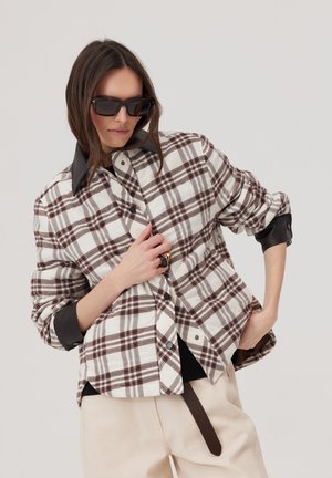 Kobieta nosząca za dużą brązowo-białą koszulę w kratę z czarnym kołnierzem i mankietami, beżowe spodnie, czarne okulary przeciwsłoneczne oraz duży pierścionek, pozująca z rękami na biodrach.