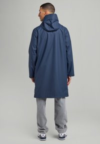 Navy raincoat with a hood, long design, waterproof material, and a relaxed fit. Worn over light gray sweatpants and sneakers.
