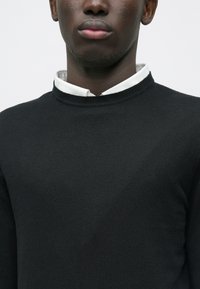 Jersey negro de punto con cuello alto y mangas largas, superpuesto a una camisa blanca con cuello visible. Textura suave.