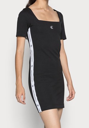 Musta lyhythihainen mekko, jossa neliskulmainen pääntie ja valkoiset sivuraidat, joissa on teksti "Calvin Klein Jeans", naisen yllä.
