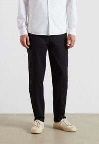 Zwarte broek van geweven stof, rechte snit, gecombineerd met beige sneakers. Witte blouse met knopen voor een deel zichtbaar aan de bovenkant.