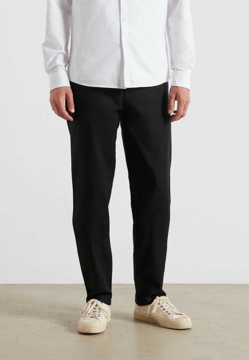 Zwarte broek van geweven stof, rechte snit, gecombineerd met beige sneakers. Witte blouse met knopen voor een deel zichtbaar aan de bovenkant.