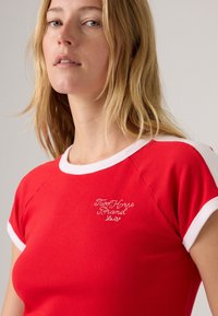 T-shirt en coton rouge avec des manches raglan blanches et un col rond. Présente un logo brodé "Two Horse Brand" sur la poitrine.
